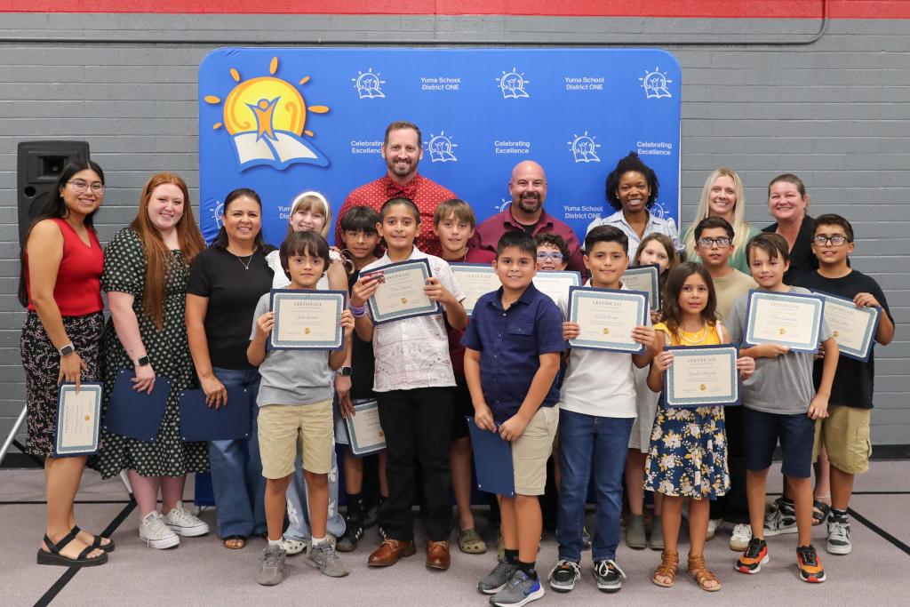A group of children and adults pose together indoors, holding certificates in front of a blue banner.